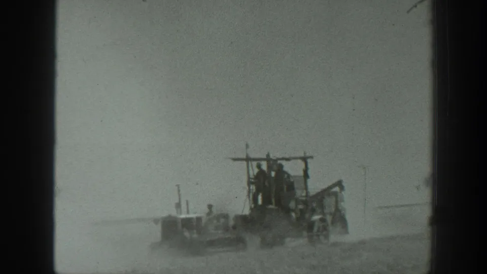 A distant 1939 California farm machine moves through dust with workers visible on an exposed platform and nearby implement sections.