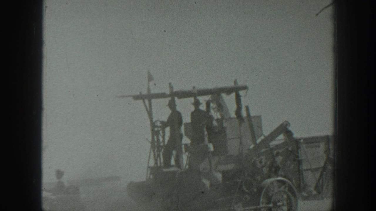 Men stand on a farm implement behind a tractor in a dusty field.