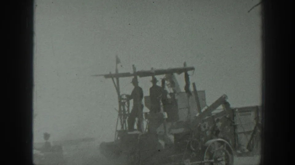 In 1939 California home-movie footage, several men appear on an open farm machine towed behind a tractor in a hazy field.