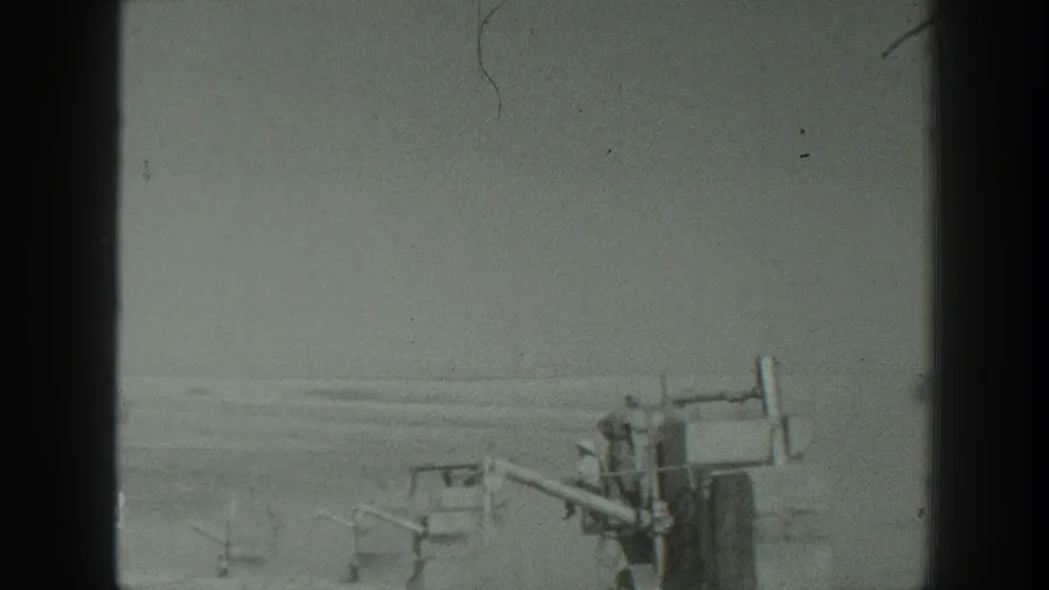 1939 California 8mm frame showing military field equipment and several figures on a broad dusty area with low hills in the distance.