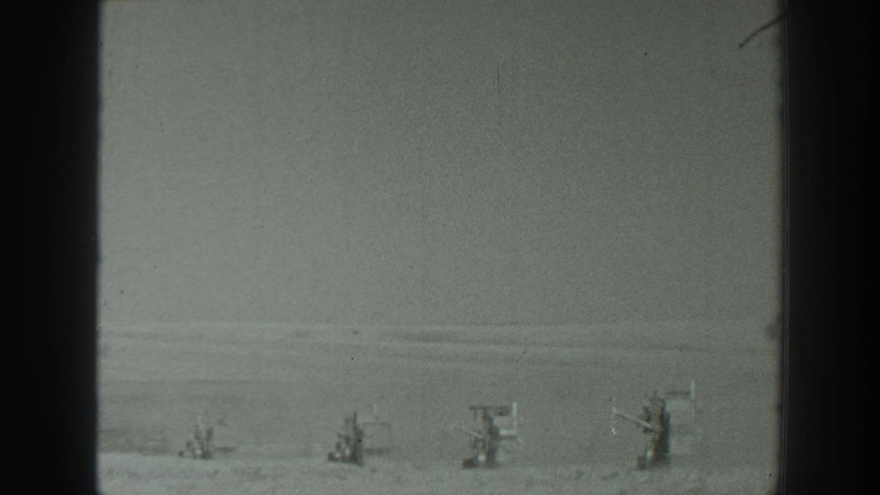 1939 California 8mm frame showing distant equipment-like structures across a flat, open landscape with soft focus and visible gate edges.