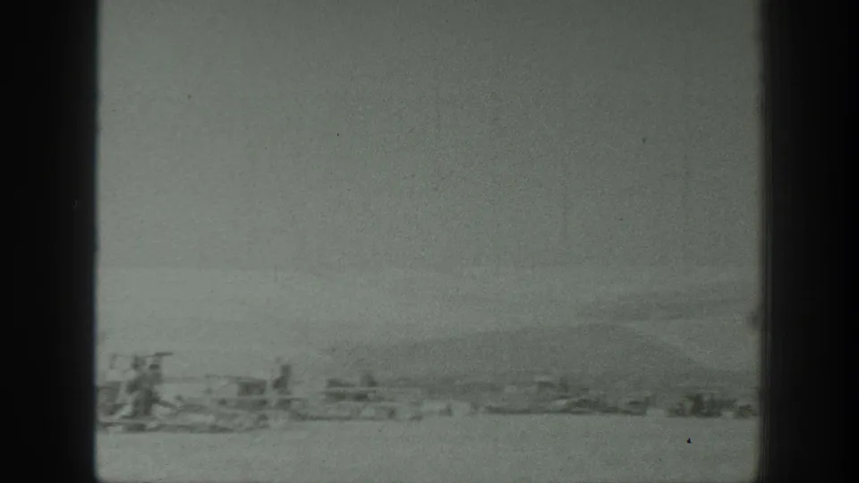 Soft black-and-white 8mm view of distant farm equipment in a California agricultural landscape, with a low horizon beyond the field.