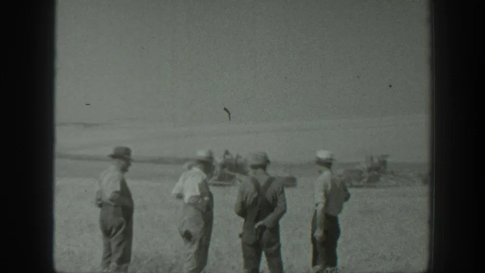 Four men in a rural California field stand near the camera while distant machinery or a vehicle sits beyond them.
