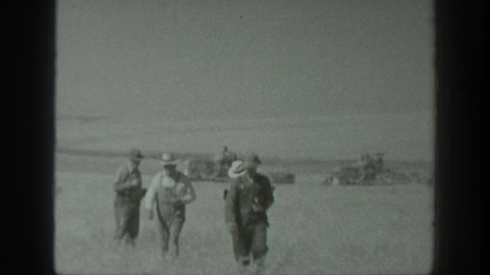 1939 California farm workers appear to walk toward the camera across a dusty field as harvesting equipment operates behind them.