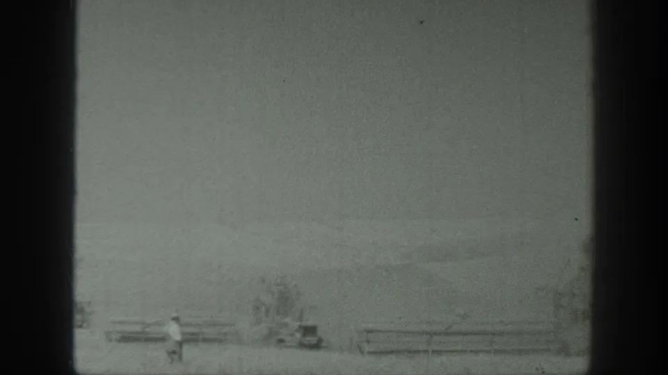 1939 California 8mm frame showing a small human figure near a fence or rail line with low farm equipment and sparse brush in the foreground field.