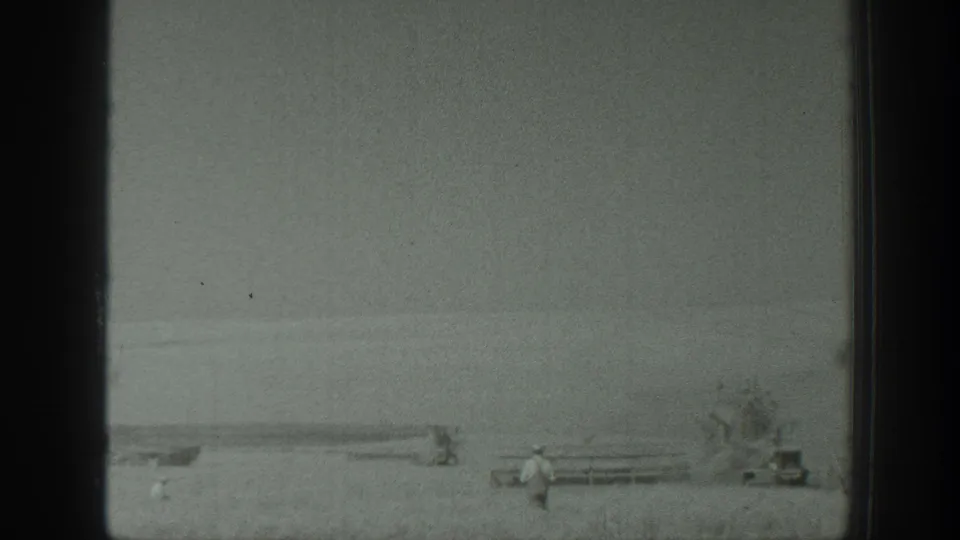 1939 California 8mm frame showing a person walking or standing near a fence or rail line, with low farm equipment, bare vegetation, and an open field beyond.