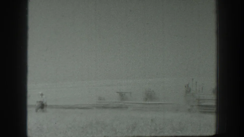 1939 California 8mm frame with a small man walking through an open field near distant vehicles or structures.