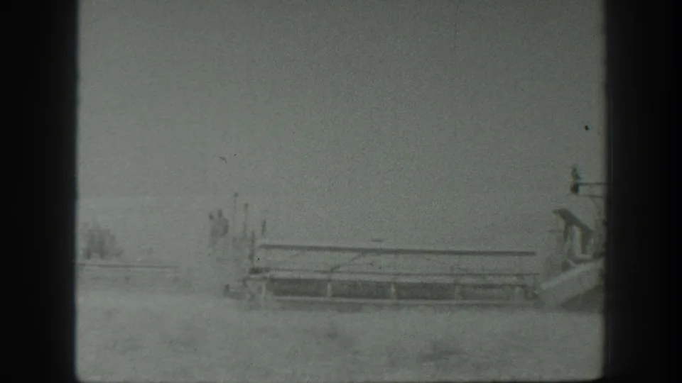 1939 California home-movie frame showing a tractor-drawn combine crossing a dusty grain field with workers faintly visible.