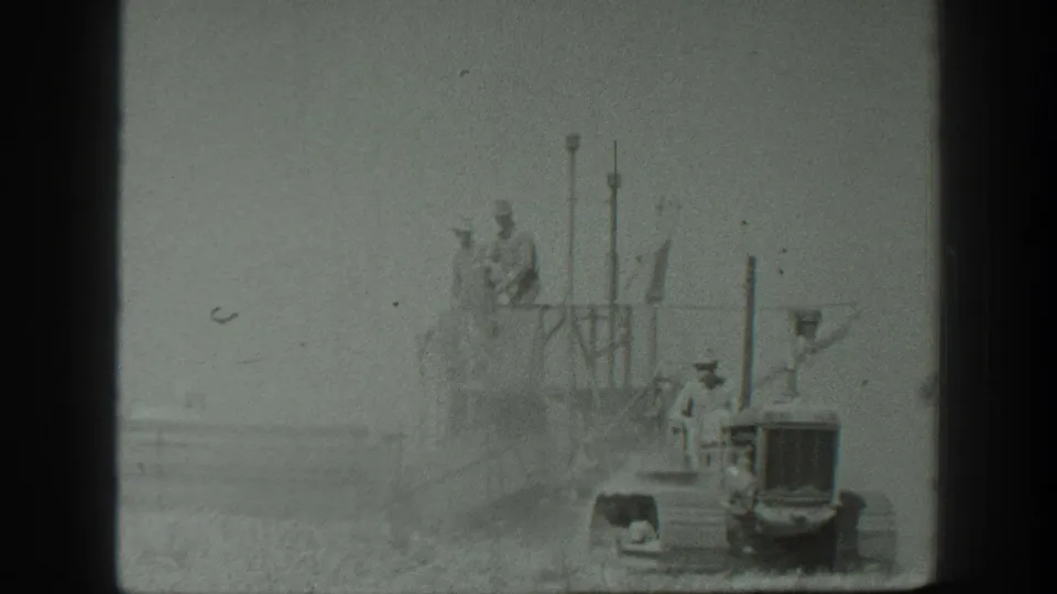 A closer 1939 California farm view shows workers riding on harvesting equipment behind a tractor in a grain field.