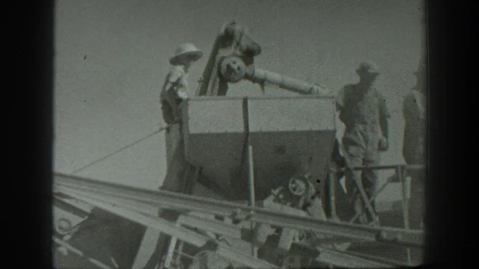 Men operate or inspect a large threshing machine in a California field, with rails or chutes visible across the foreground.