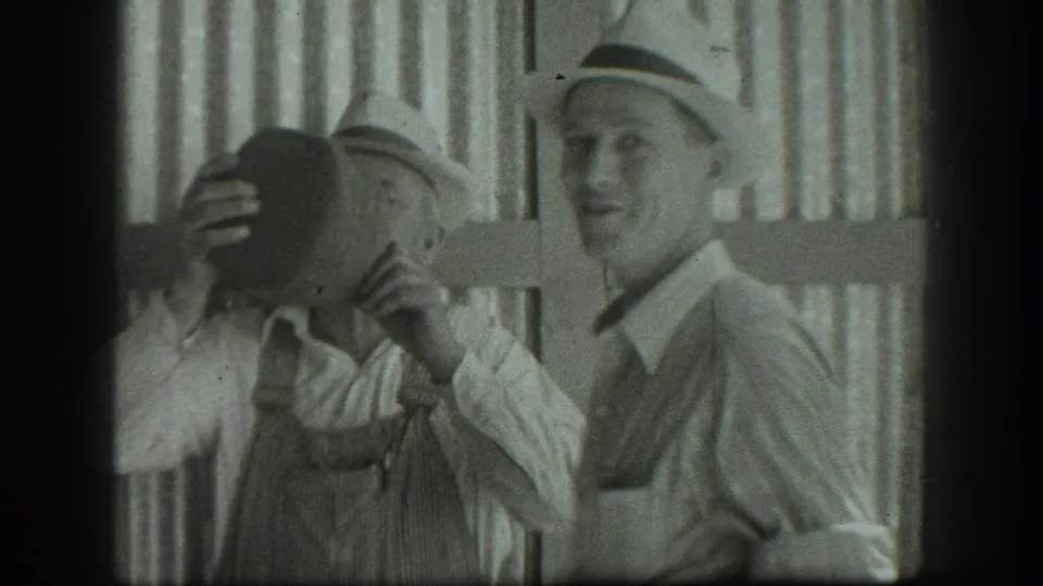 Two adult men in hats appear outdoors in 1939 California, one lifting a large jug while the other smiles beside wooden siding.