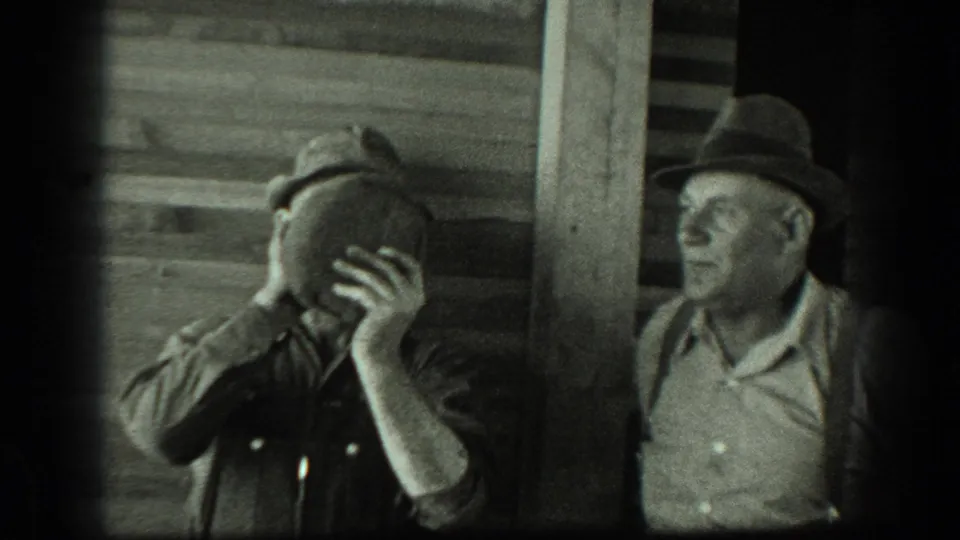 In a black-and-white 1939 California frame, one man drinks from a jug as another hatted man watches beside a rustic wooden wall.