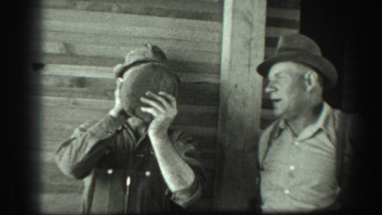 Two men stand by a wooden wall as one raises a canteen to his face.