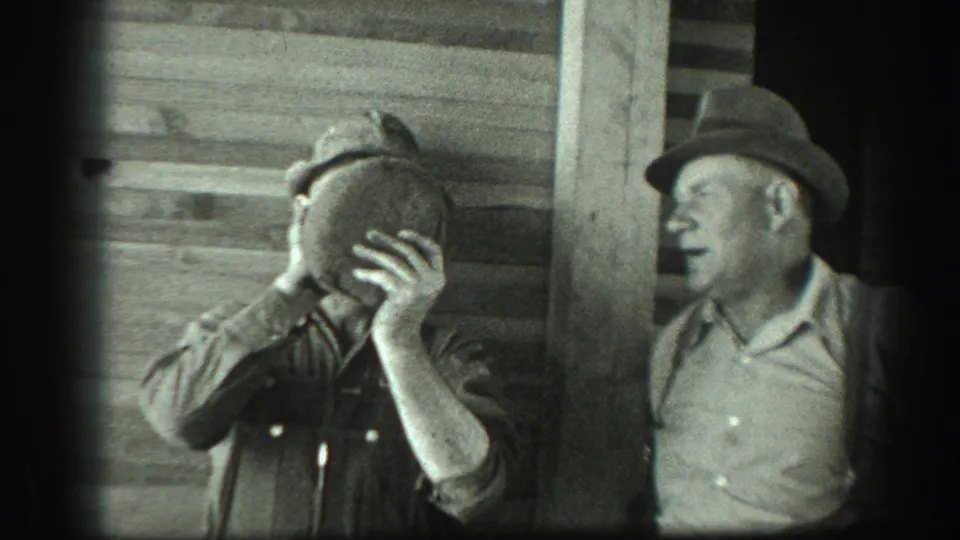 In 1939 California home-movie footage, two adult men stand beside rough wooden siding while one lifts a canteen and the other watches.
