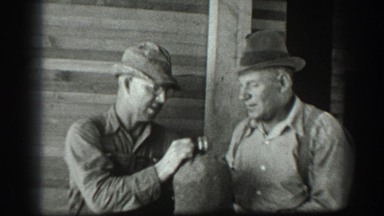 A man in a cap offers or passes a canteen toward a man in a hat.