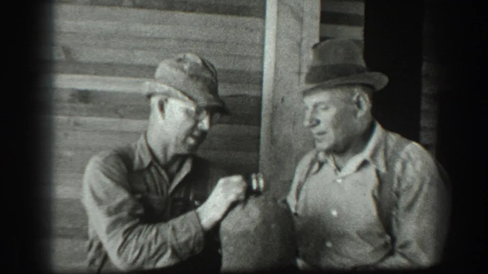 Two adult men in work clothes stand against wooden siding as the man in a cap extends the canteen toward the man in a fedora.