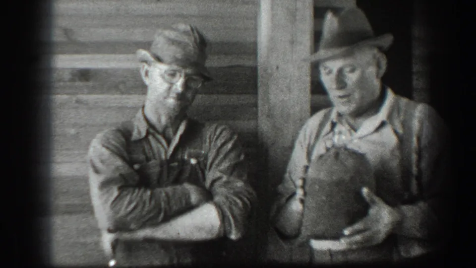 The man in overalls stands with folded arms as the other man in suspenders holds the canteen in front of the wooden structure.