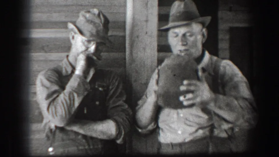 In the final frame, the man in a fedora drinks from the canteen while the man in a cap stands nearby against the wooden wall.