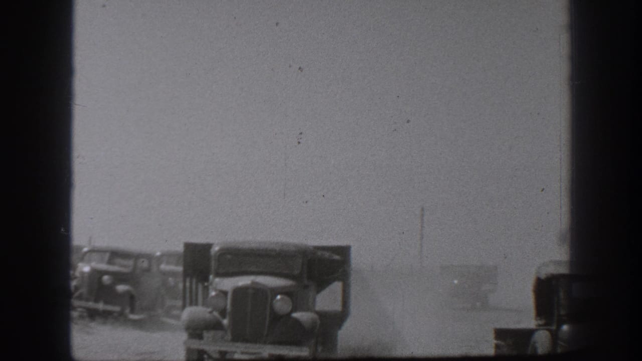 Several boxy trucks line a dusty open area with a dark foreground post.