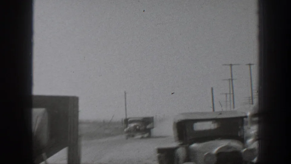 Wide 1939 California rural road view with vintage vehicles traveling in dust, utility poles along the roadside, and a partially framed vehicle body in front.