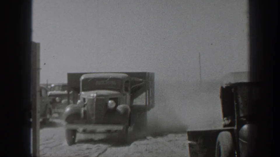 1939 California 8mm frame showing a vintage truck on a dusty road, with another vehicle at frame edge, background automobiles, utility poles, and open rural land.