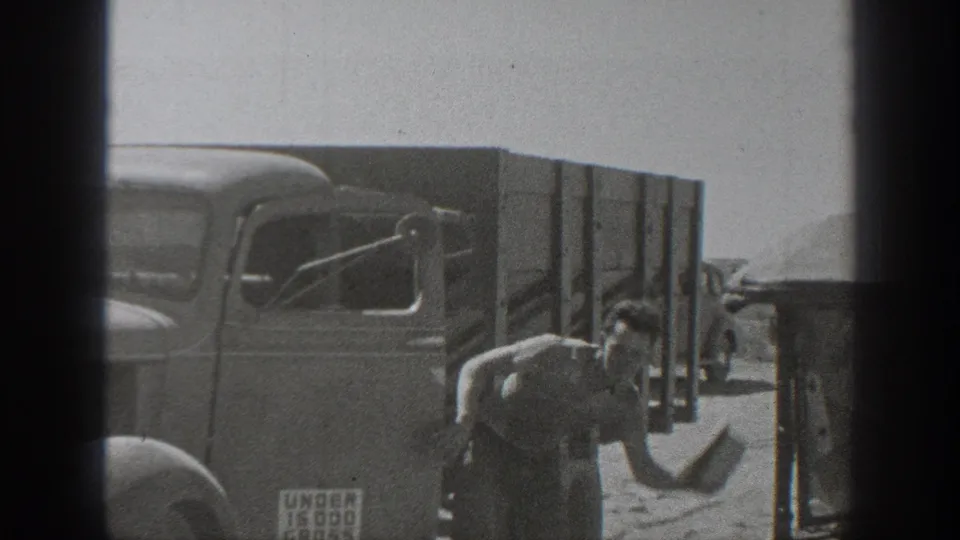 A worker stands near a 1930s-style truck in California, with dust, a load sign, and roadside activity visible.
