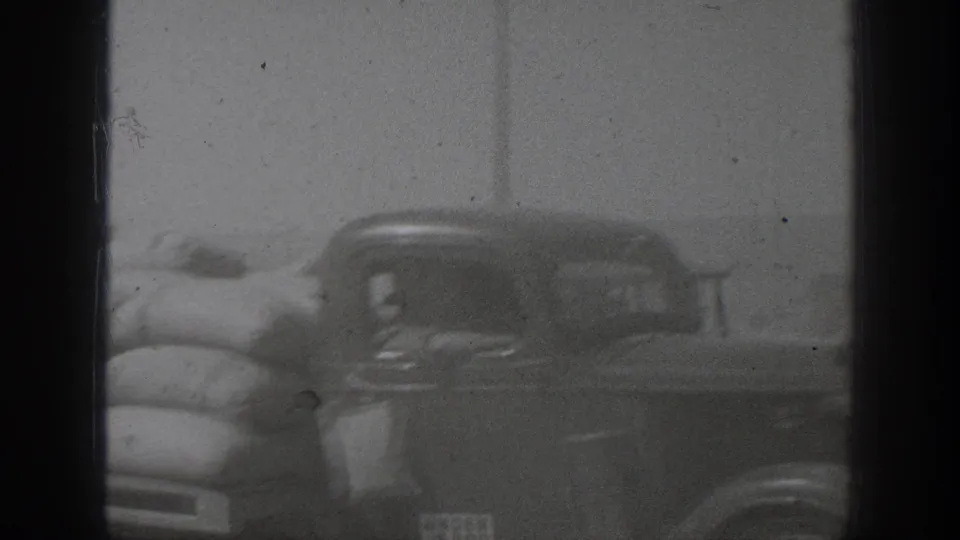 Soft black-and-white frame of a vintage car near large sacks, suggesting goods being moved or loaded.