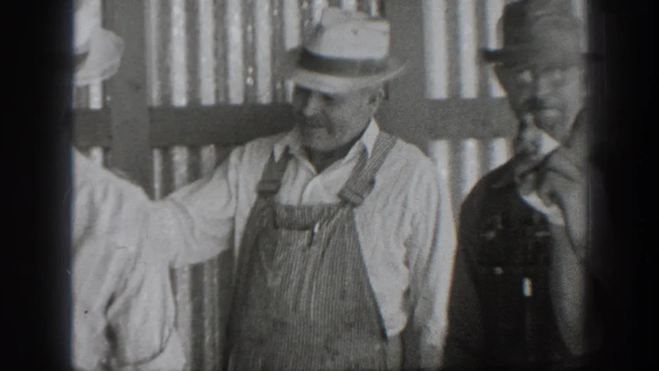 1939 California frame with a man in a striped apron and brimmed hat standing with another worker near vertical slats.