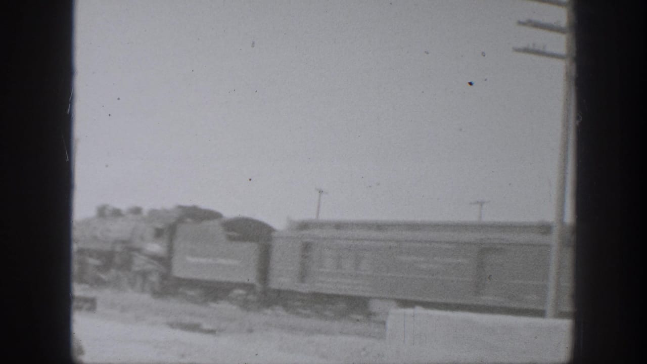 1939 California 8mm frame showing a steam locomotive pulling rail cars past open ground with utility poles in the distance.
