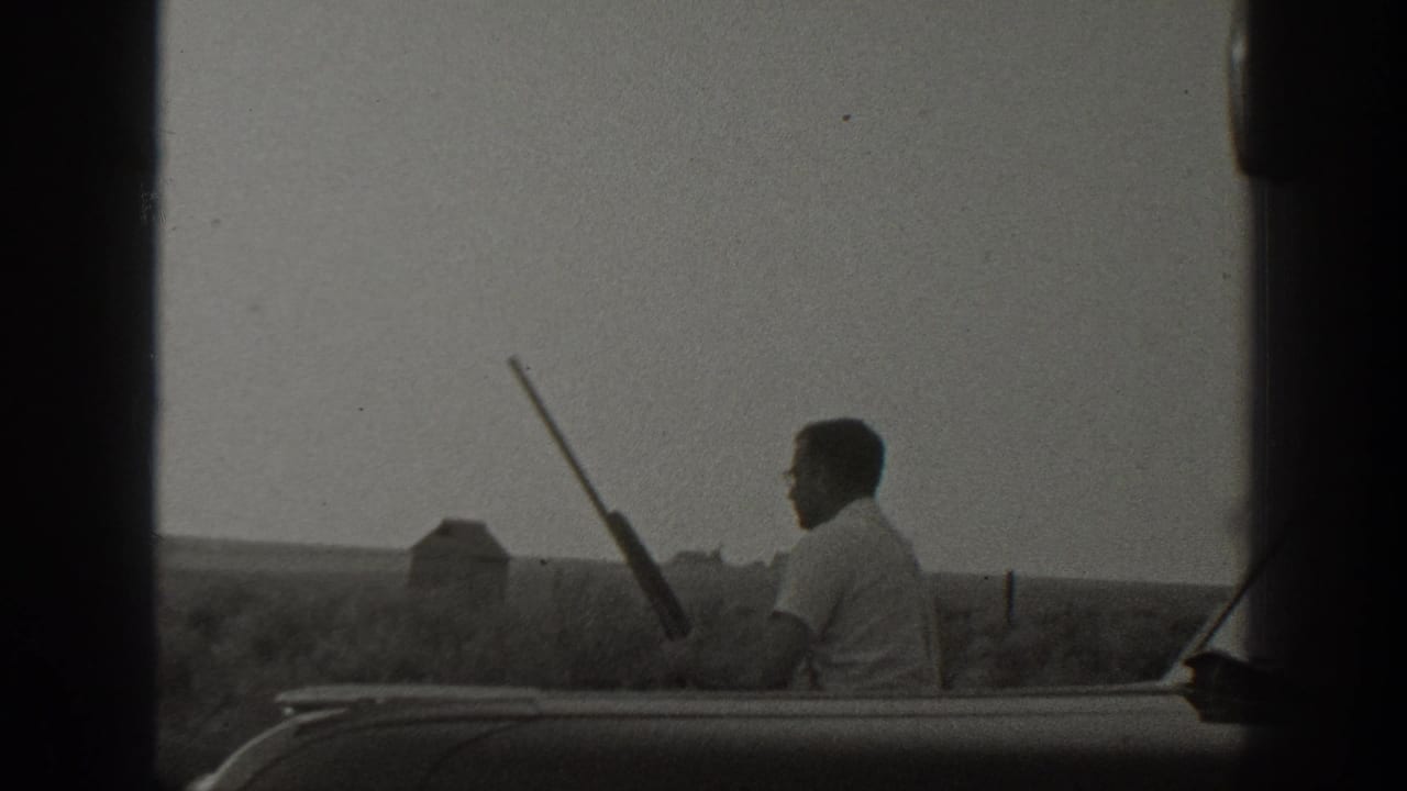 Black-and-white 1939 California home-movie frame of a man seated at a car edge holding a rifle in a rural field setting.