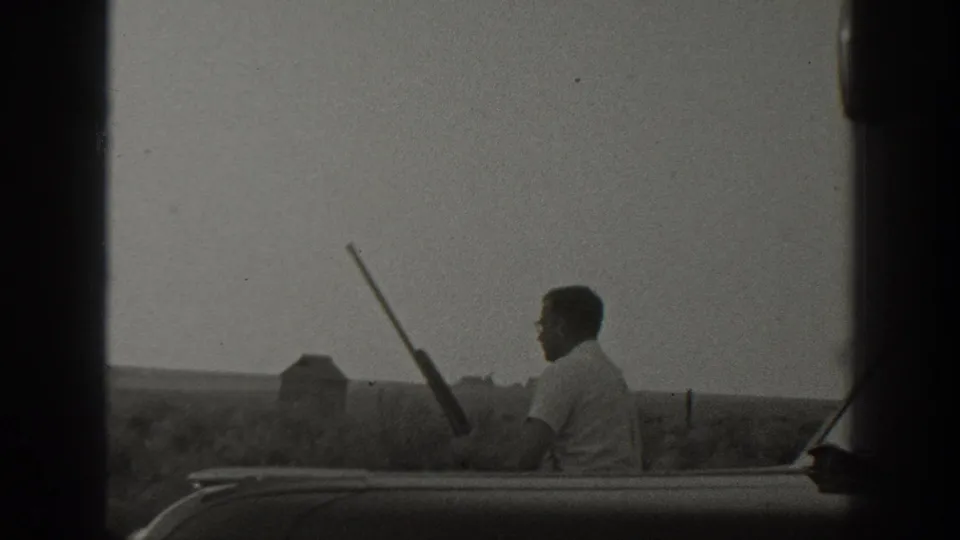 Black-and-white 1939 California home-movie frame of a man seated at a car edge holding a rifle in a rural field setting.