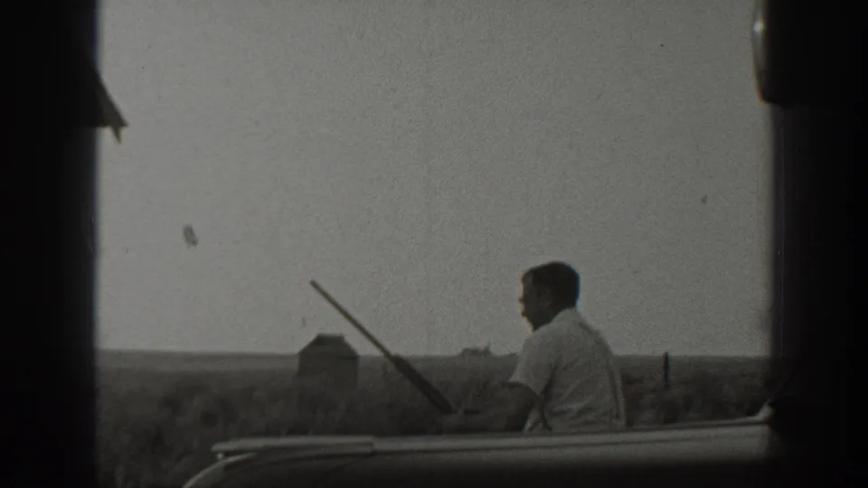 1939 California 8mm still showing a seated adult man with a rifle near a car, set against open rural fields.