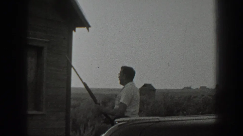 Black-and-white 1939 California frame of a man with a rifle near a car, with a small wooden structure at the left edge.