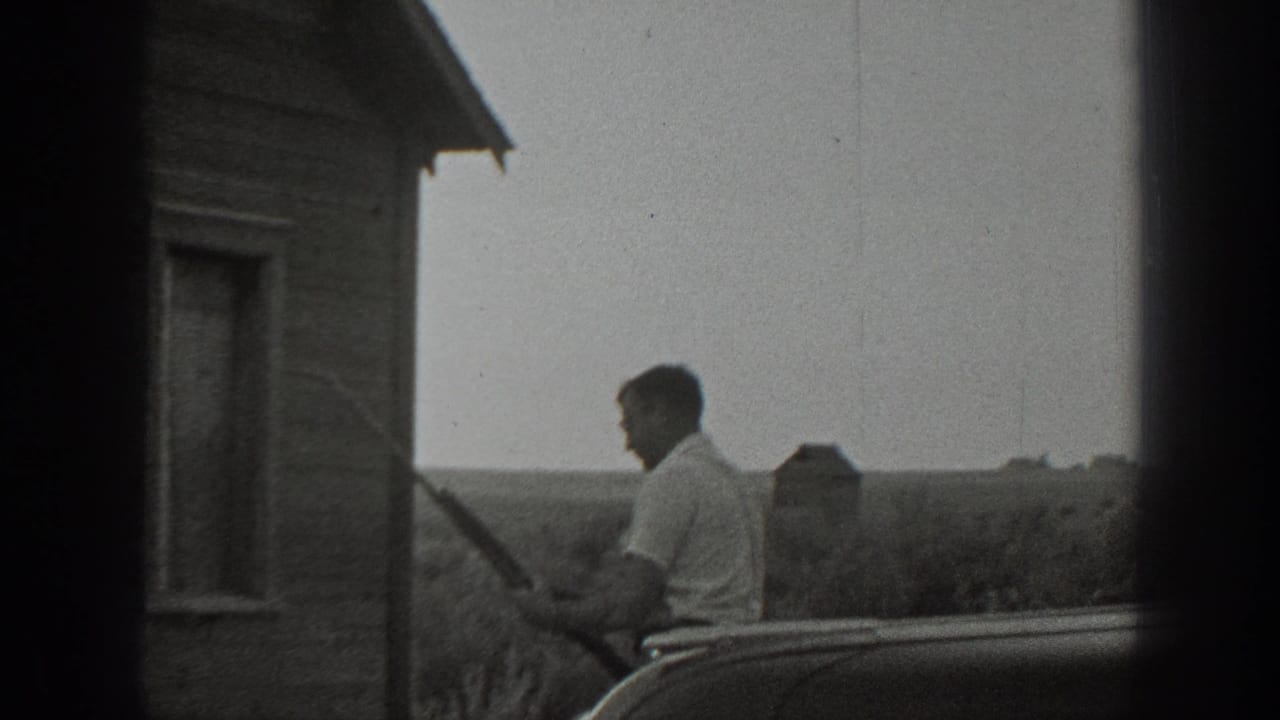 In 1939 California home-movie footage, a man stands near a wood-sided rural house and appears to hold a rifle beside a partial automobile.