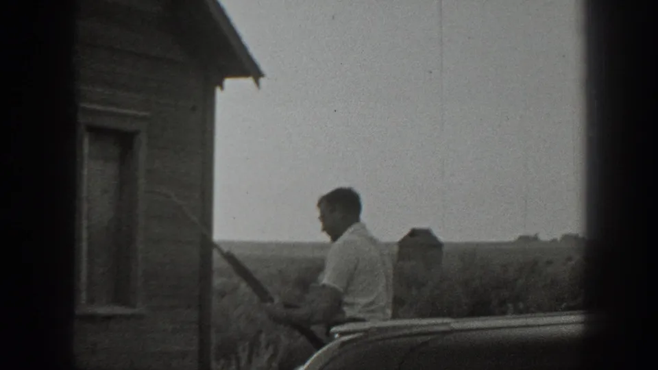 In 1939 California home-movie footage, a man stands near a wood-sided rural house and appears to hold a rifle beside a partial automobile.