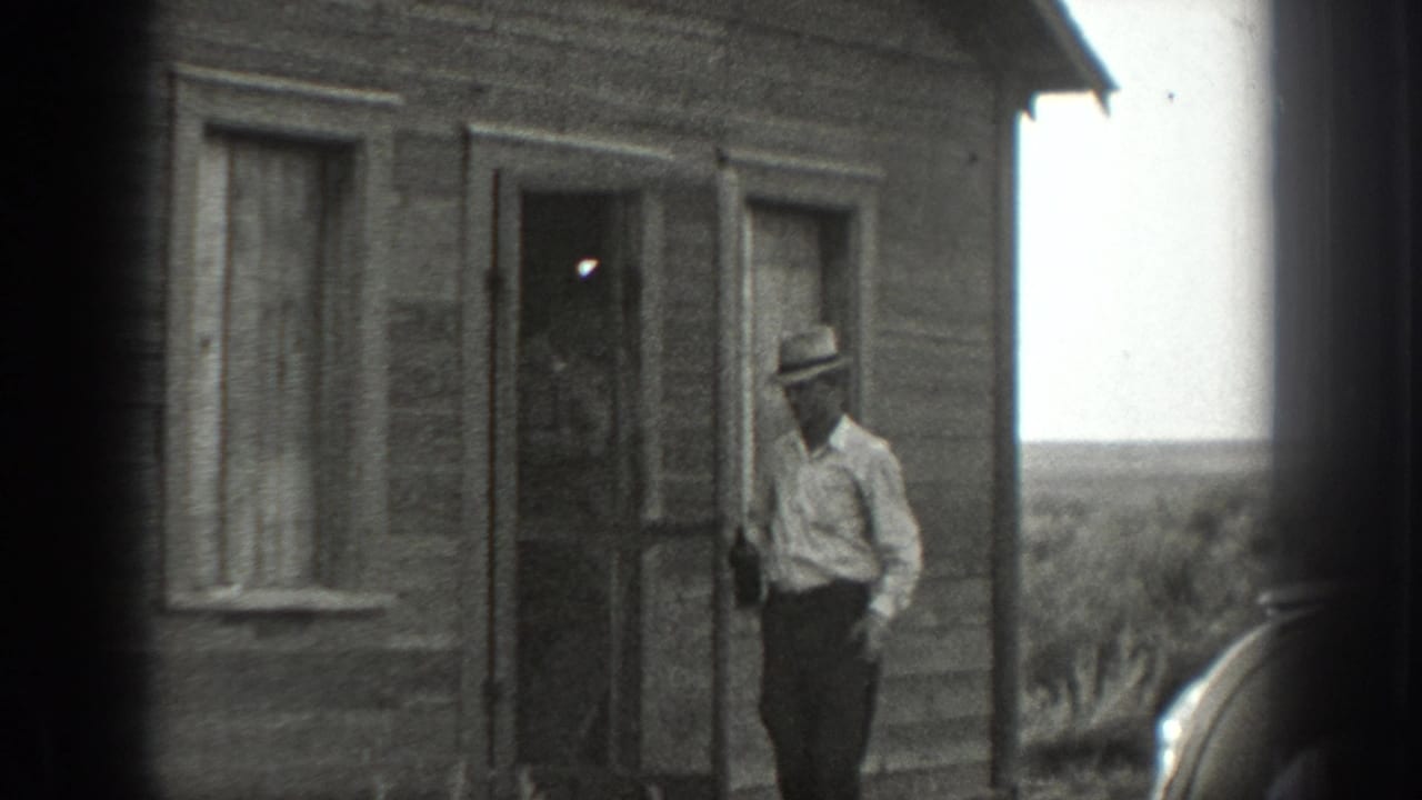 A man in a light shirt and hat stands near the doorway of a weathered rural wooden house, with open land visible beyond.