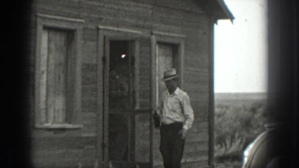 A man in a light shirt and hat stands near the doorway of a weathered rural wooden house, with open land visible beyond.
