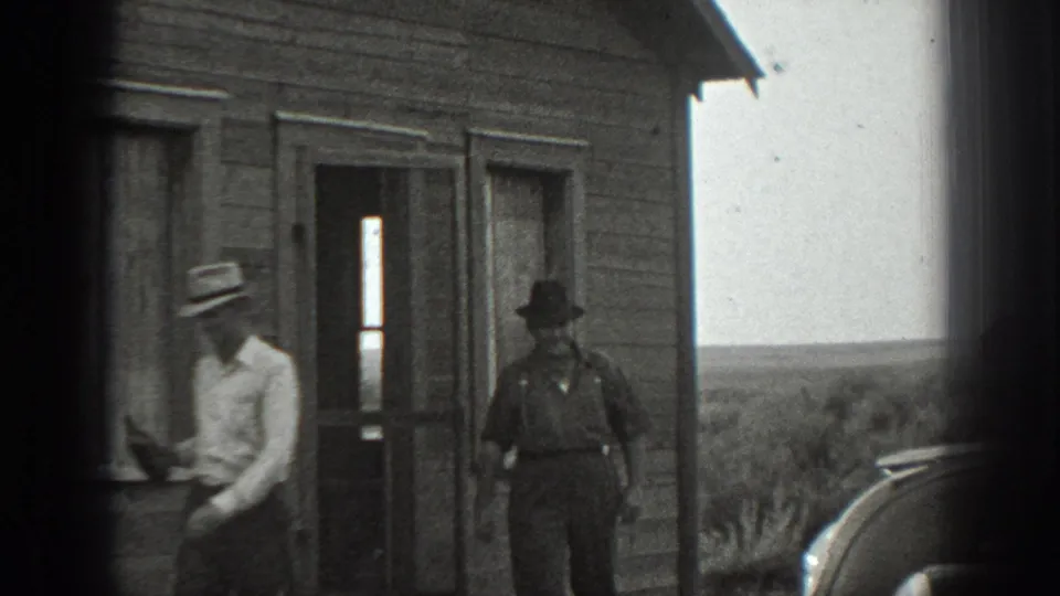 Two adult men are outside a rural wooden house in the supplied 1939 California footage, one near the wall and another by the doorway with a rifle.