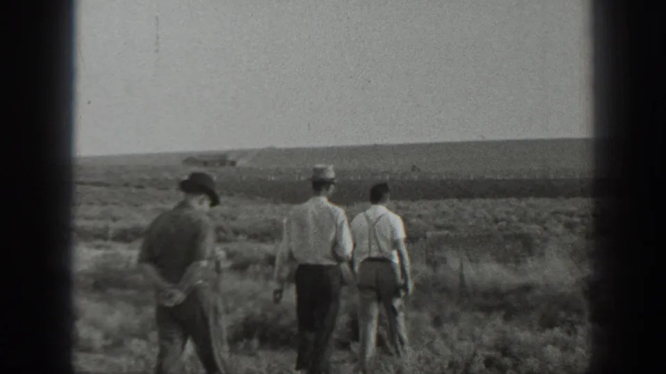 1939 California home-movie frame with three men in light shirts and hats walking through scrubland, one carrying a firearm at his side.