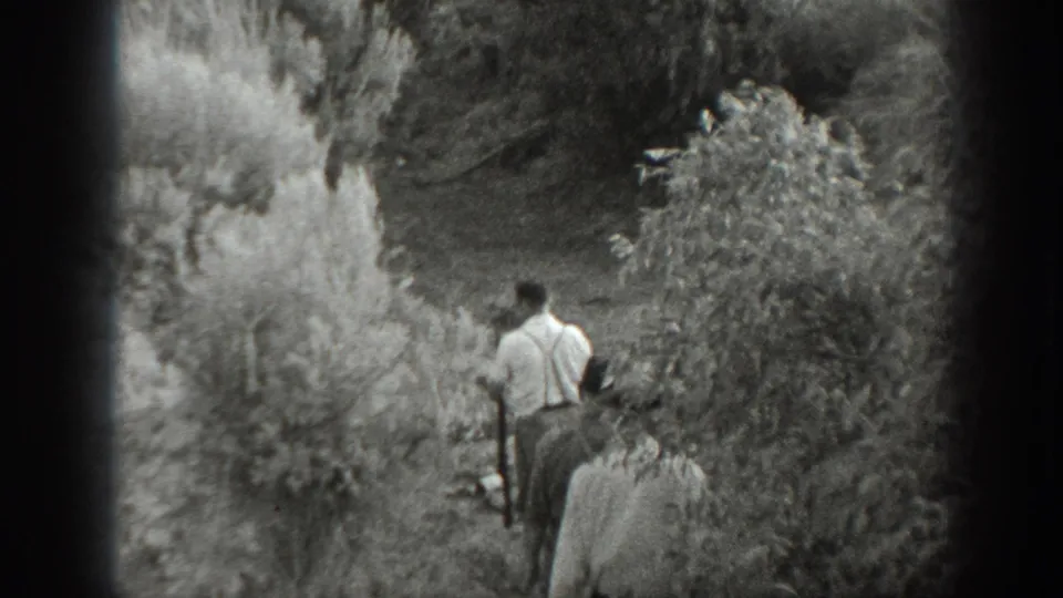 1939 California home-movie frame of men moving along a narrow path through dense vegetation, with suspenders and period clothing visible.