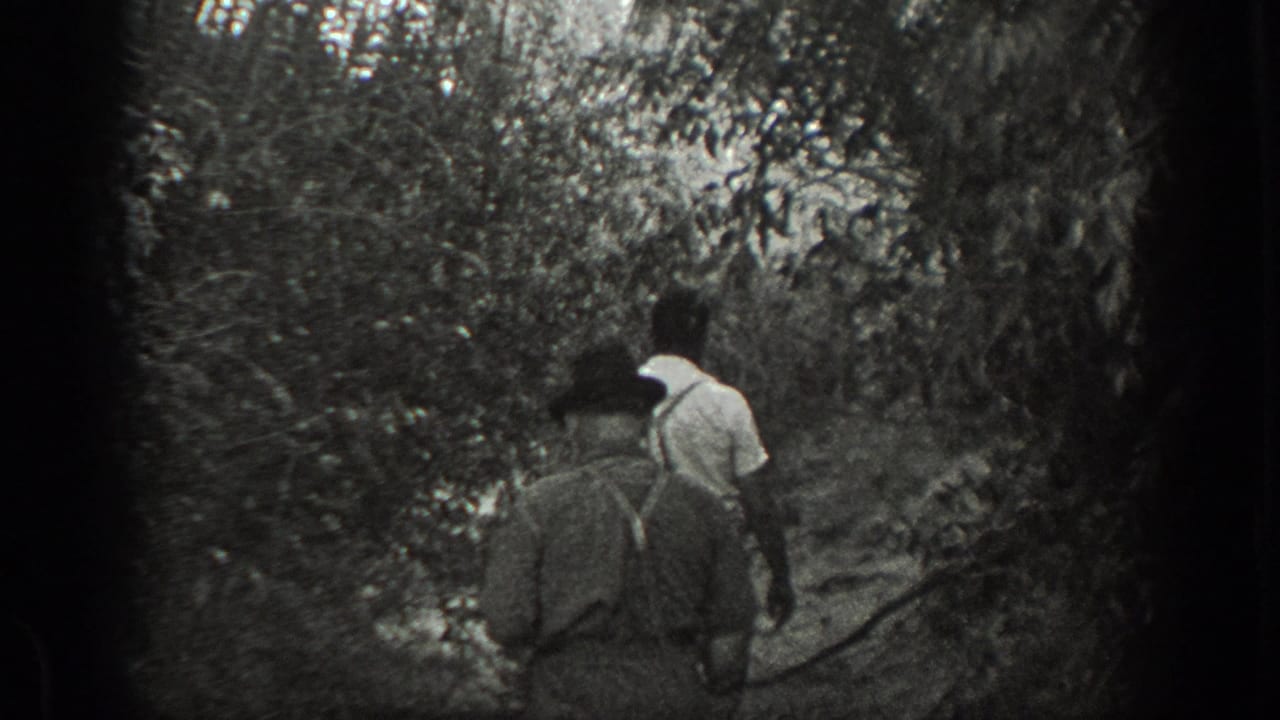 Two men walk away along a narrow path through dense trees.
