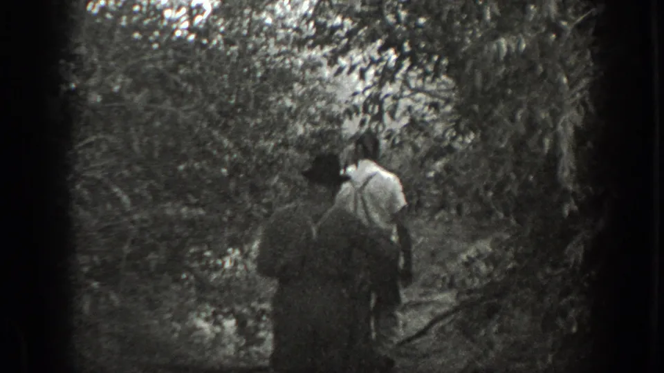 Two men walk deeper into dense California woodland, filmed from behind on black-and-white 8mm.