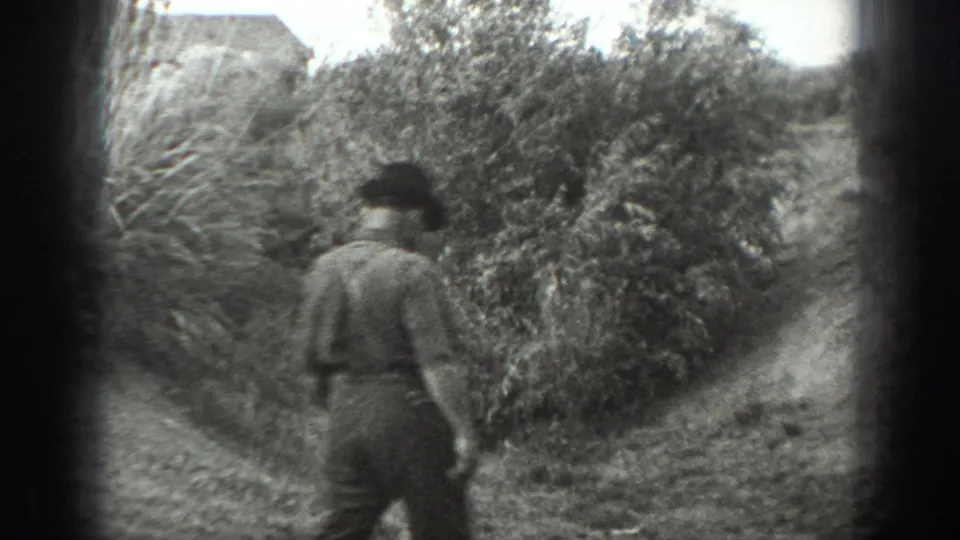 A single adult man walks away along a dirt path beside thick brush in black-and-white 8mm California footage.