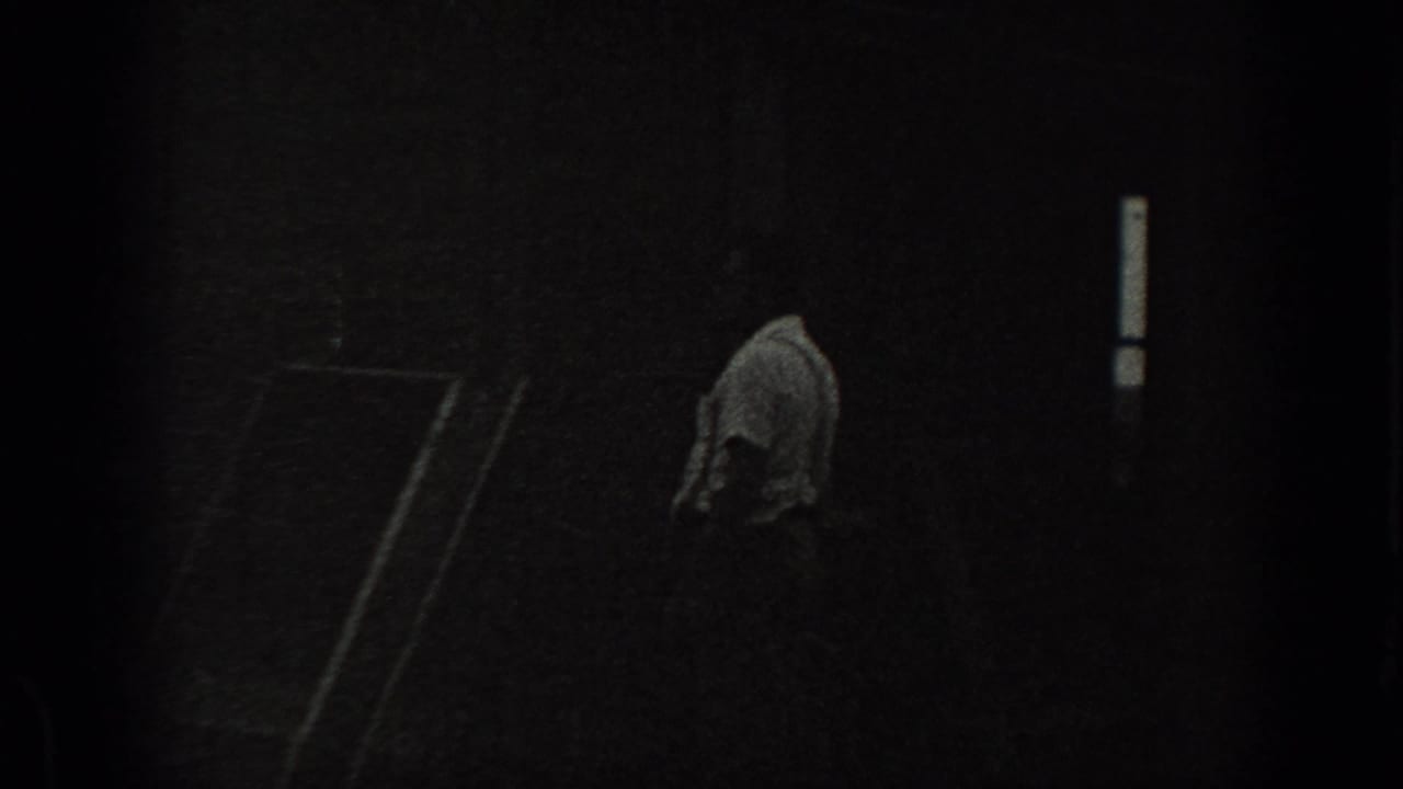 A man stands or walks near a dark house wall with a narrow bright window.