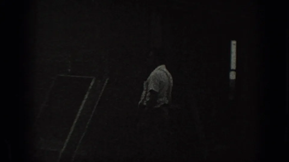 A man in a light shirt appears near the dark exterior wall of a rural house, with a small bright window visible.