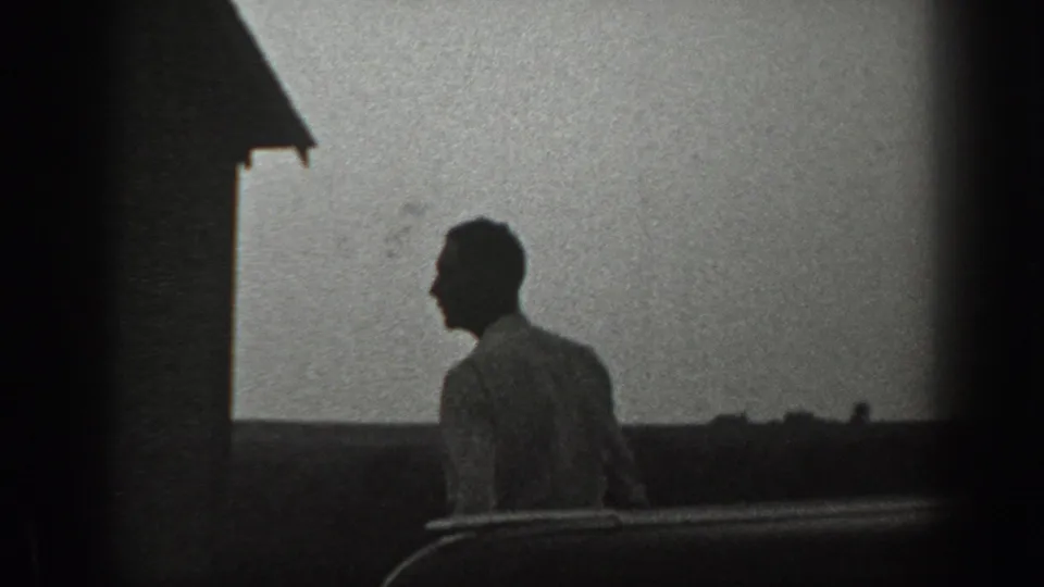 A man in profile moves past a building edge against a pale sky in black-and-white 8mm footage.