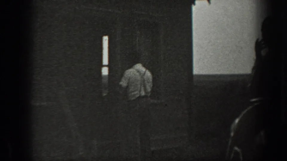 A man in a light shirt moves beside a dark rural house wall, with a bright vertical window and pale sky beyond.