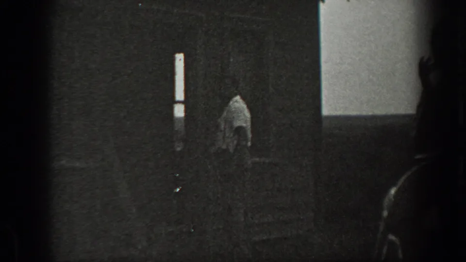 1939 California home-movie frame showing a dim interior, a man near a vertical window, and another shadowed figure partly visible at right.