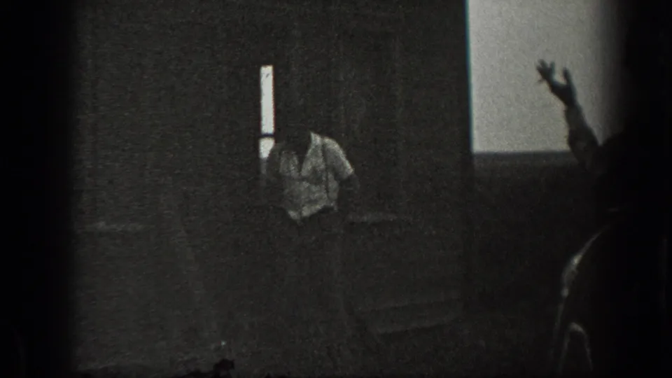 Dim 1939 California interior frame with a light-shirted man near the wall and a partially visible person gesturing in silhouette.