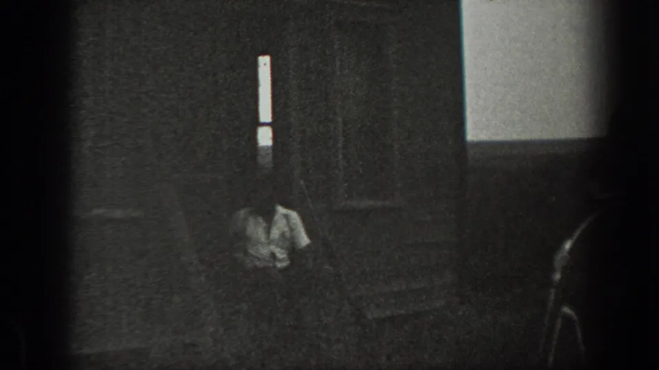 Grainy 1939 California home-movie frame showing a man crouched or seated low in a dark room beneath a narrow vertical window.